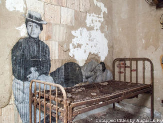 French Artist, JR, depicts blown up photos of immigrants on the wall in front of an old-fashioned bed in the Ellis Island Hospital