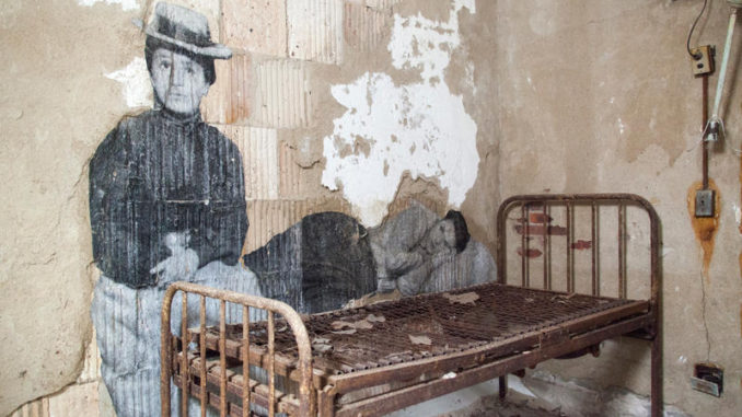 French Artist, JR, depicts blown up photos of immigrants on the wall in front of an old-fashioned bed in the Ellis Island Hospital