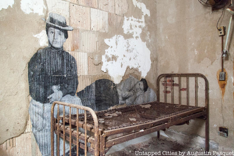 French Artist, JR, depicts blown up photos of immigrants on the wall in front of an old-fashioned bed in the Ellis Island Hospital
